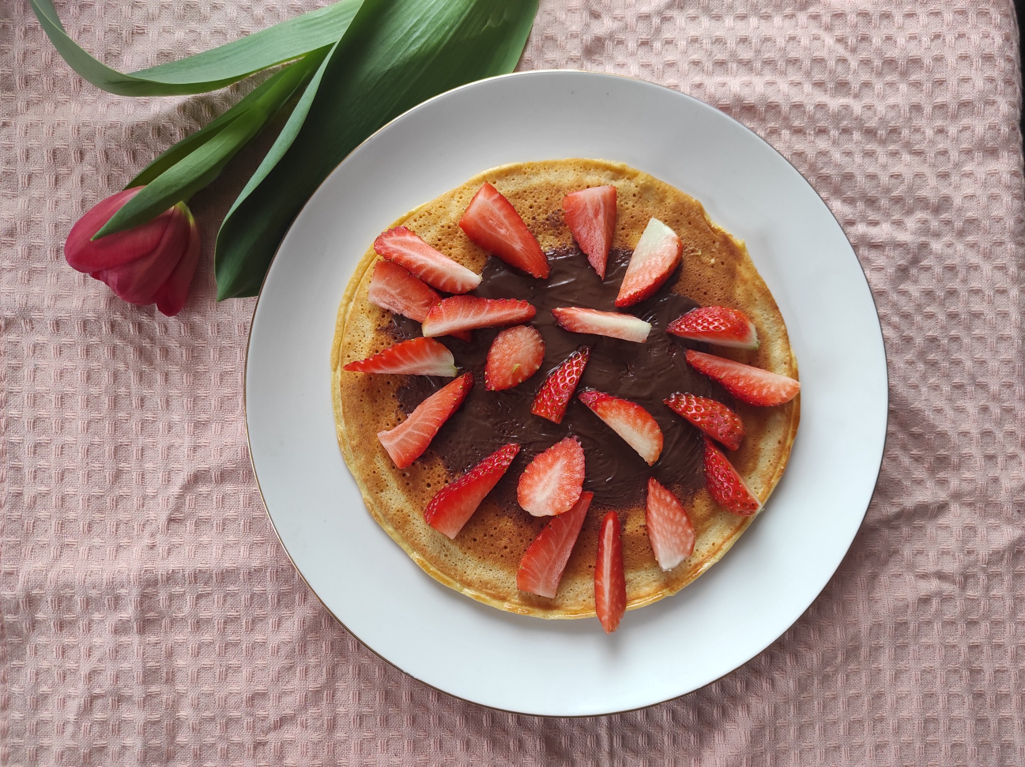 Puszysty naleśnik polany kremem czekoladowym i udekorowany plasterkami świeżych truskawek, podany na białym talerzu z tulipanem na różowej teksturowanej ściereczce.