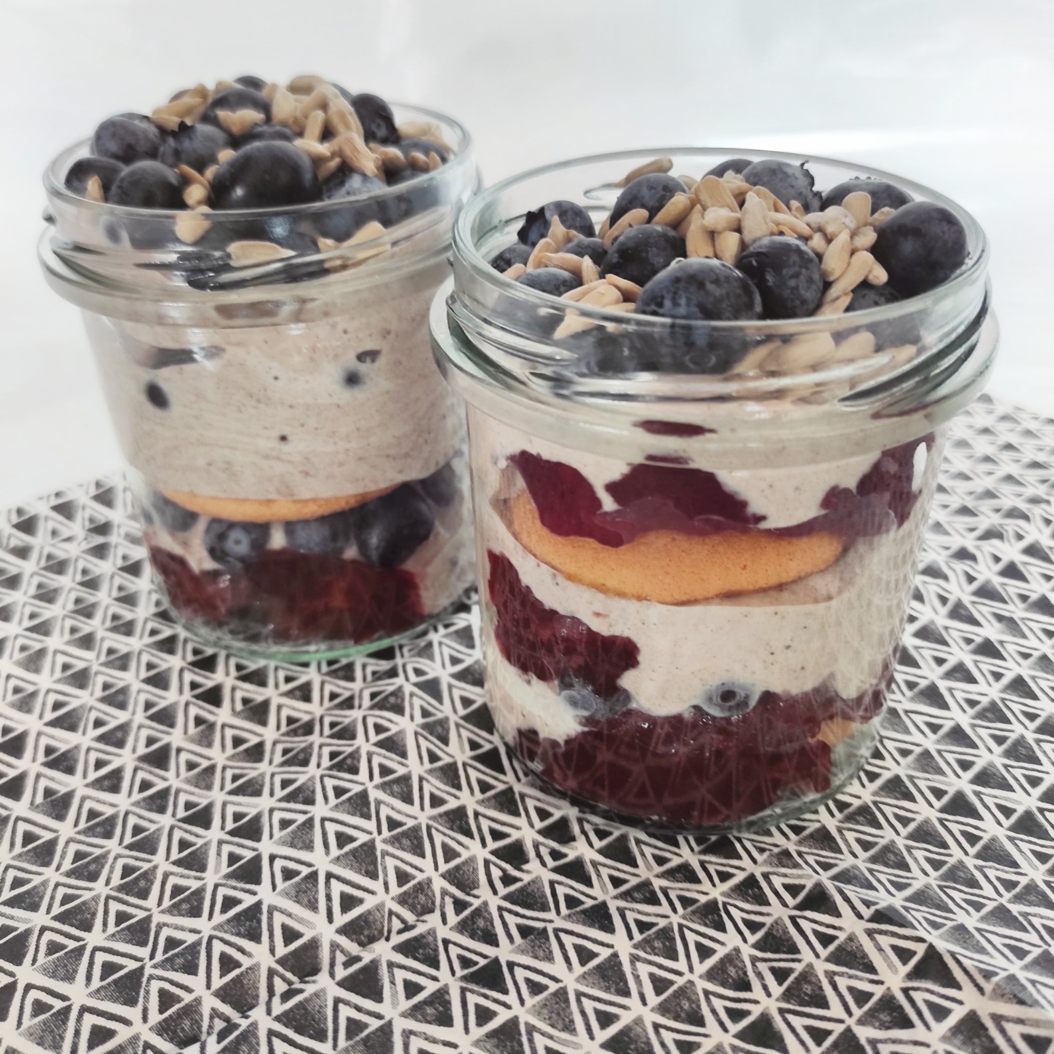 Dwa słoiki z deserem warstwowym: na dnie wiśnie, krem, biszkopt i na wierzchu borówki z ziarnami słonecznika, ustawione na geometrycznej podkładce.