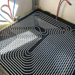 Budotom Pompy Ciepła - Białe rury ogrzewania podłogowego ułożone na czarnej macie izolacyjnej w rogu pomieszczenia z szarymi ścianami i wystającymi rurami instalacji wodnej.