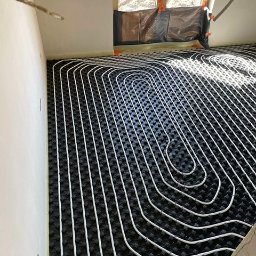 Budotom Pompy Ciepła - Białe rury ogrzewania podłogowego ułożone na czarnej macie izolacyjnej w jasnym pomieszczeniu z oknem zabezpieczonym folią. Widoczne detale instalacji.