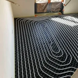 Białe rury ogrzewania podłogowego ułożone na czarnej macie izolacyjnej w jasnym pomieszczeniu z oknem zabezpieczonym folią. Widoczne detale instalacji.