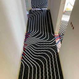 Budotom Pompy Ciepła - Rozłożona instalacja ogrzewania podłogowego na czarnej macie izolacyjnej, z widocznymi białymi rurami i czerwono-niebieskimi złączkami w korytarzu w trakcie budowy.