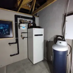 Biała pompa ciepła Panasonic Aquarea z czarnymi rurami na szarej ścianie. Widoczny zmiękczacz wody i drewniana konstrukcja stropu. Szare płytki podłogowe.