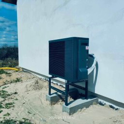 Budotom Pompy Ciepła - Nowoczesna pompa ciepła typu monoblok, zainstalowana na zewnątrz budynku na betonowym fundamencie. Widoczne przyłącza i fragment elewacji w surowym stanie.