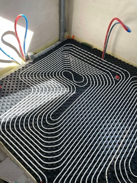 Białe rury ogrzewania podłogowego ułożone na czarnej macie izolacyjnej w rogu pomieszczenia z szarymi ścianami i wystającymi rurami instalacji wodnej.