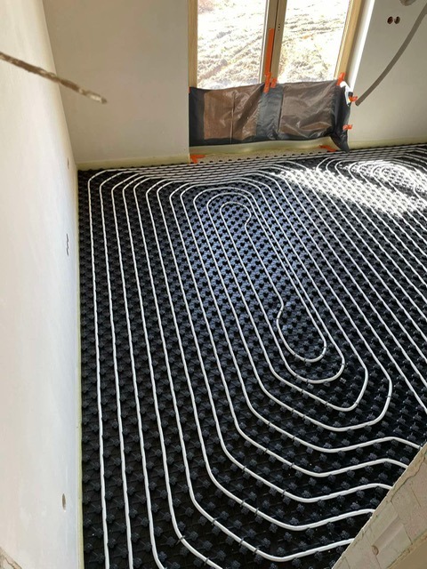 Białe rury ogrzewania podłogowego ułożone na czarnej macie izolacyjnej w jasnym pomieszczeniu z oknem zabezpieczonym folią. Widoczne detale instalacji.