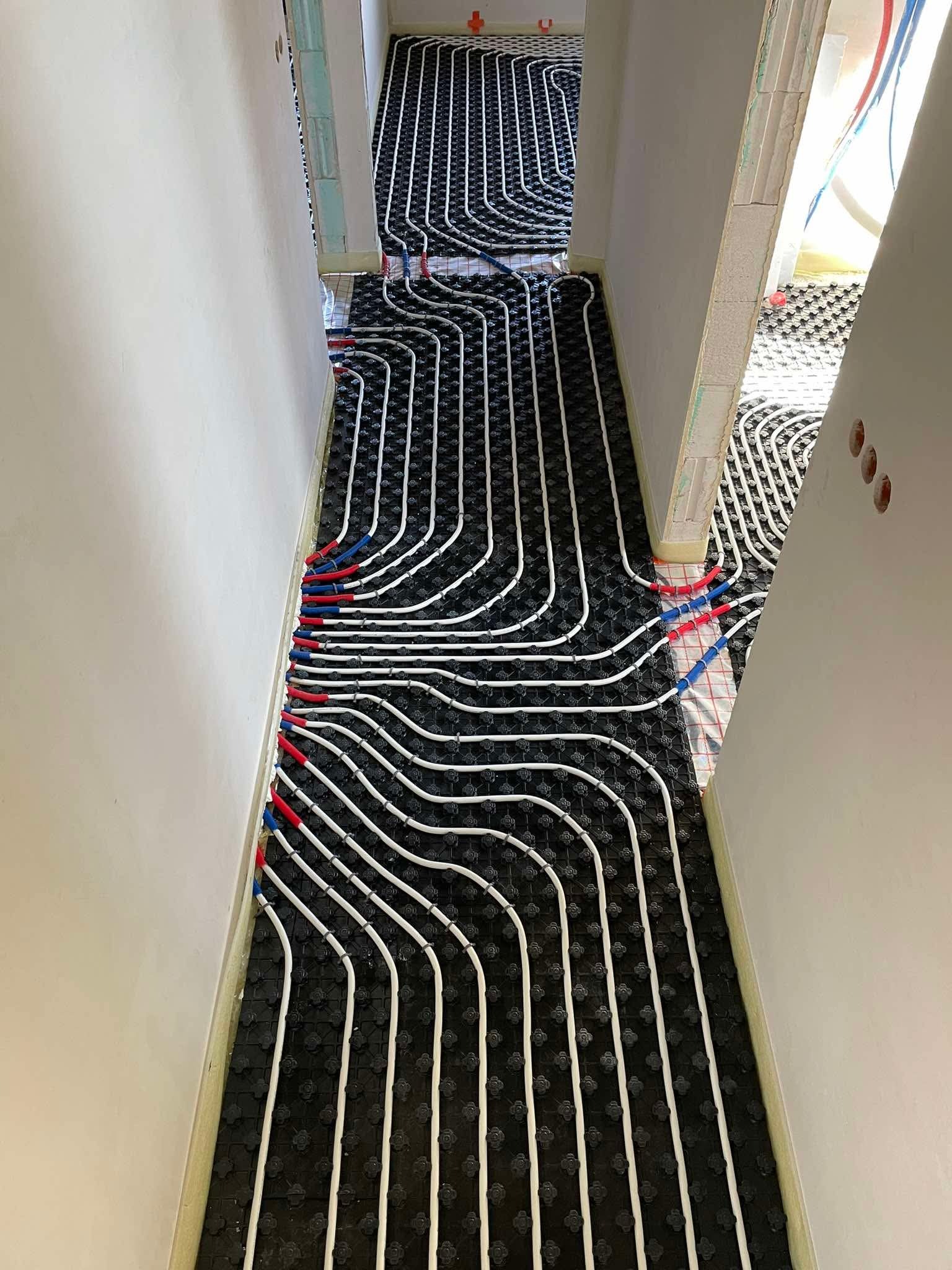 Rozłożona instalacja ogrzewania podłogowego na czarnej macie izolacyjnej, z widocznymi białymi rurami i czerwono-niebieskimi złączkami w korytarzu w trakcie budowy.