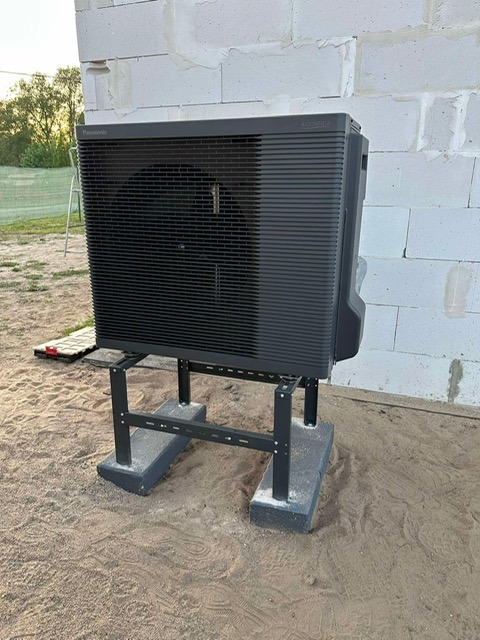 Szara pompa ciepła Aquarea na czarnym stelażu, ustawiona na betonowych blokach, przy ścianie z białych bloczków. Widoczne ślady stóp na piasku.