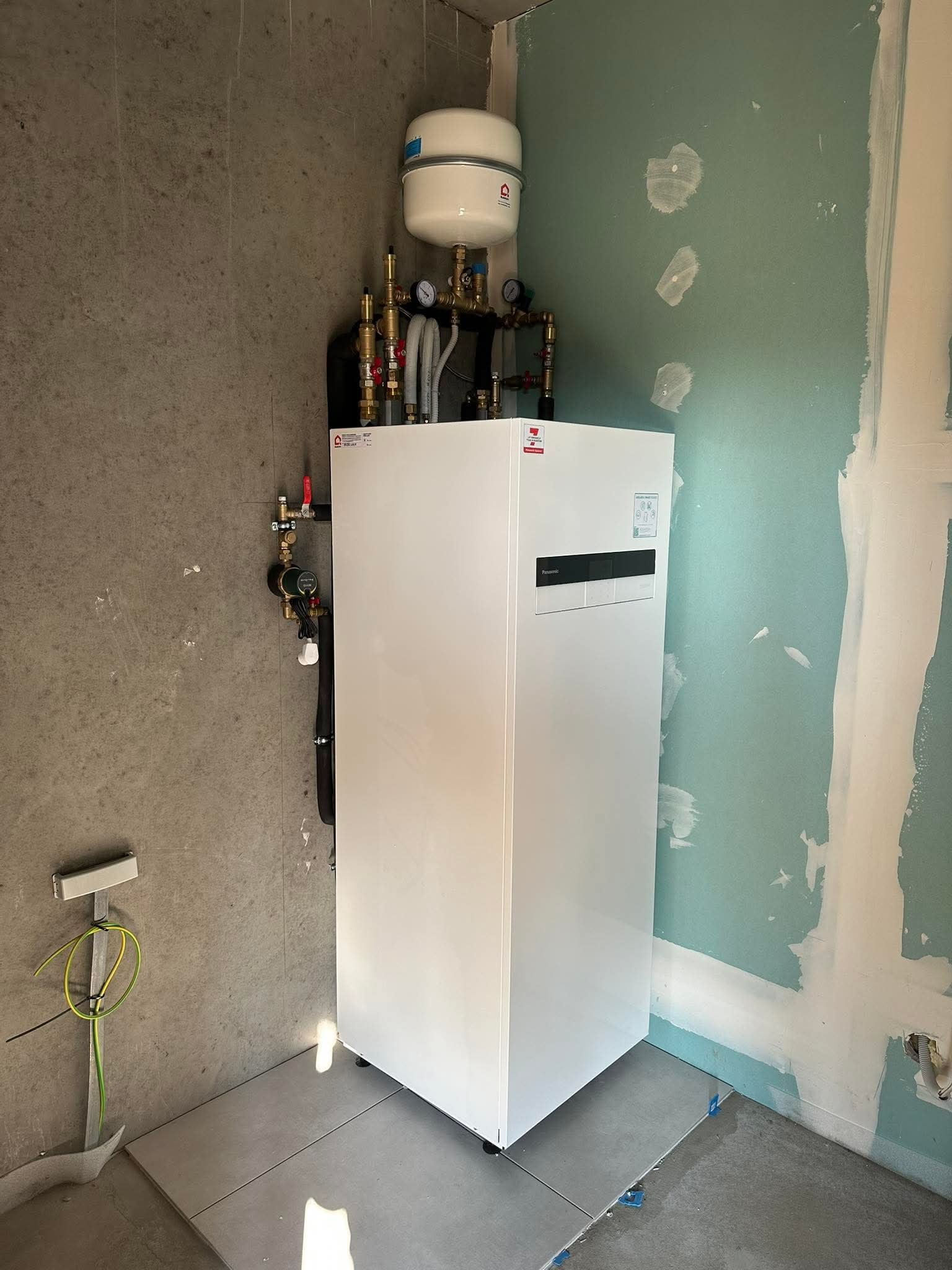 Biała pompa ciepła Panasonic z podłączoną instalacją, na tle surowych ścian i częściowo wyłożonej podłogi płytkami. Widoczne elementy instalacji hydraulicznej.