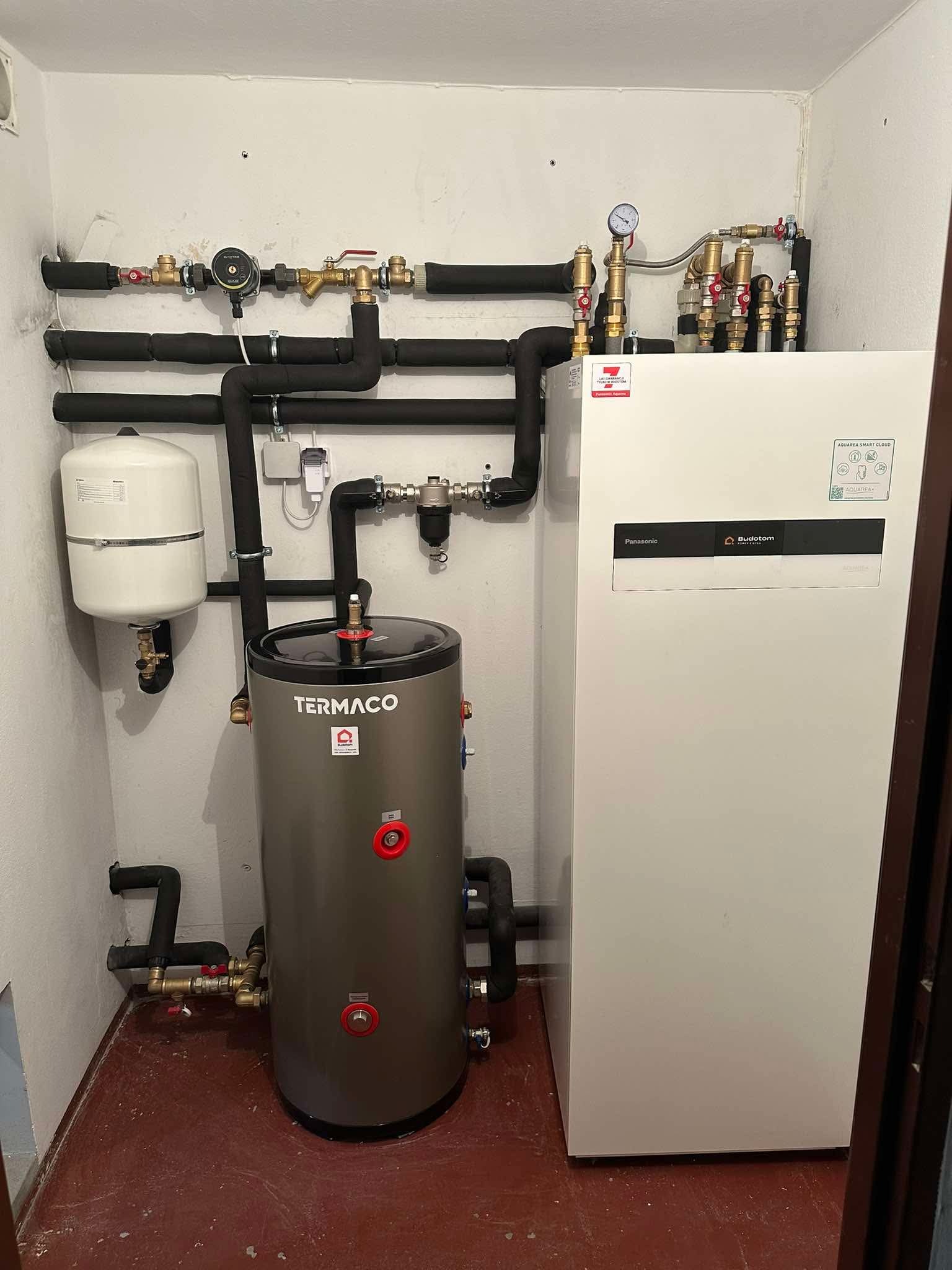 Instalacja grzewcza w kotłowni: zbiornik Termaco, pompa ciepła Panasonic Budotom, rury z izolacją, manometr. Realizacja w pomieszczeniu z czerwoną posadzką.