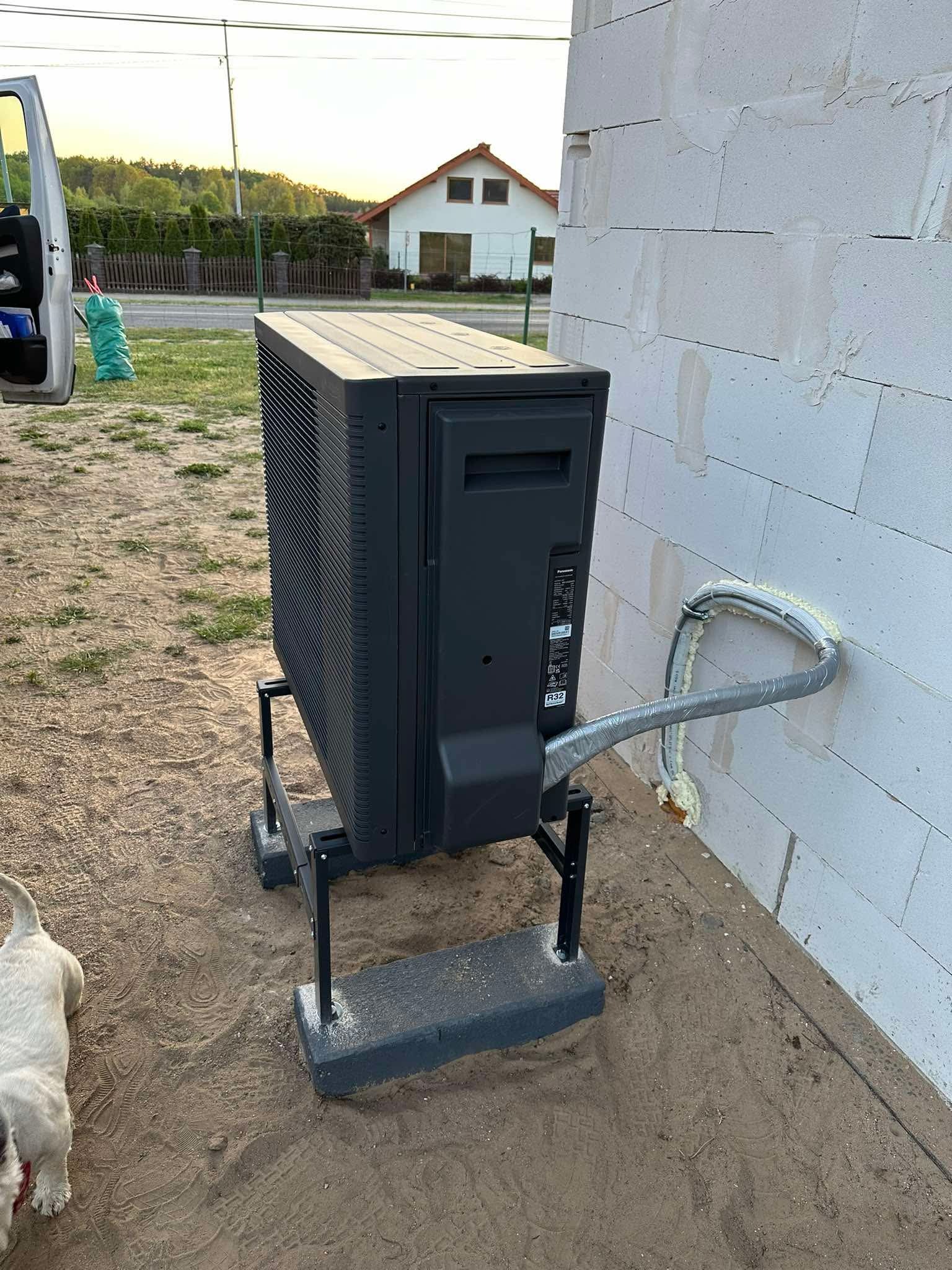 Zamontowana, zewnętrzna jednostka pompy ciepła Panasonic na metalowej konstrukcji przy ścianie z betonu komórkowego. Widoczne rury z izolacją i uszczelnieniem.