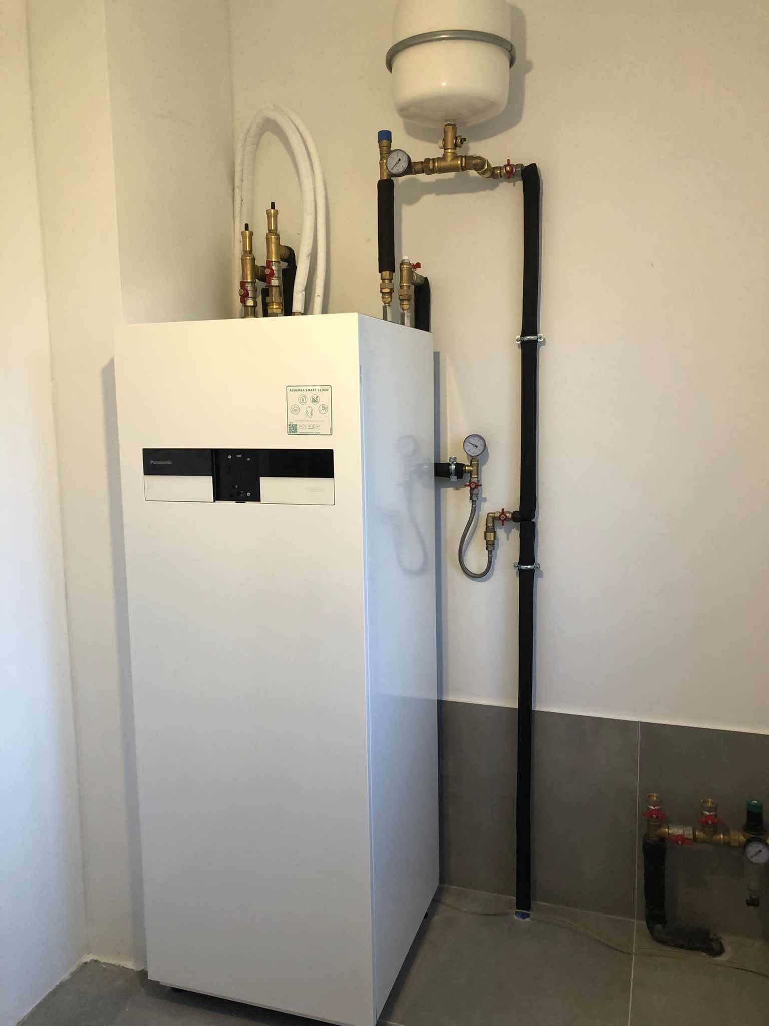 Biała pompa ciepła Panasonic Aquarea Smart Cloud z instalacją hydrauliczną i zbiornikiem wyrównawczym w jasnym pomieszczeniu z szarą glazurą.