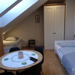 Pokój na poddaszu z dwoma łóżkami pojedynczymi, stolikiem z koronkową serwetą i pilotami, oraz szafą z białymi drzwiami.