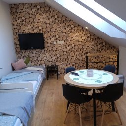 Przedsiębiorstwo Usługowe Komplex - Pokój w pensjonacie z dwoma łóżkami, okrągłym stołem z czarnymi krzesłami i ścianą wyłożoną plastrami drewna, pod skosami dachu z oknami.