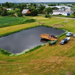 Przedsiębiorstwo Usługowe Komplex - Widok z góry na staw z drewnianym pomostem, grupą osób i zaparkowanymi pojazdami w wiejskim otoczeniu.