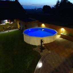 Przedsiębiorstwo Usługowe Komplex - Podświetlony, okrągły basen ogrodowy z białym obramowaniem i niebieską wodą, otoczony drewnianym tarasem i trawnikiem, oświetlony lampami wzdłuż ogrodzenia, widok wieczorny.