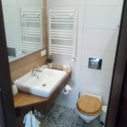 Nowoczesna łazienka z drewnianymi akcentami: blat pod umywalką, rama lustra i deska sedesowa. Białe płytki, grzejnik drabinkowy, umywalka nablatowa i wzorzysta podłoga.