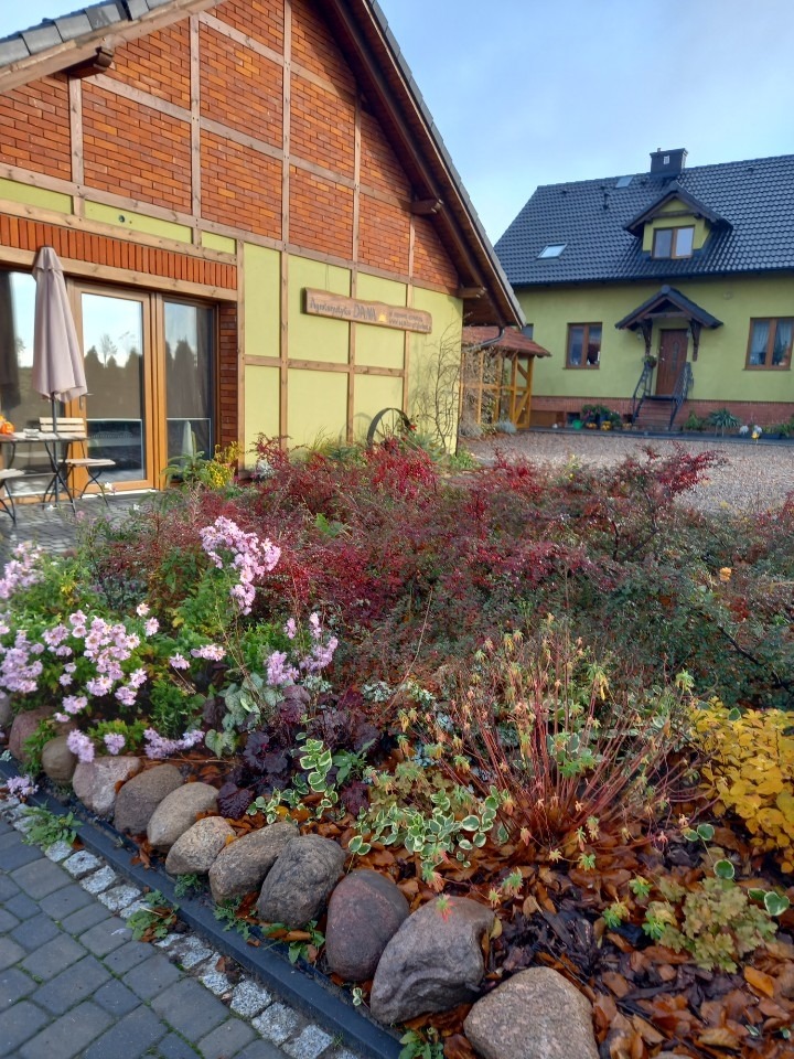 Ujęcie z zewnątrz budynku ośrodka wypoczynkowego z ogrodem, widoczny kamienny murek, rabaty z kwitnącymi kwiatami oraz budynek z tabliczką informacyjną.