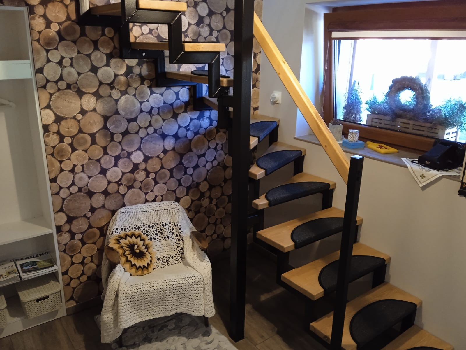 Wnętrze pokoju z drewnianą ścianą imitującą pnie drzew, krzesłem z narzutą i schodami z drewnianymi stopniami i czarną metalową konstrukcją.