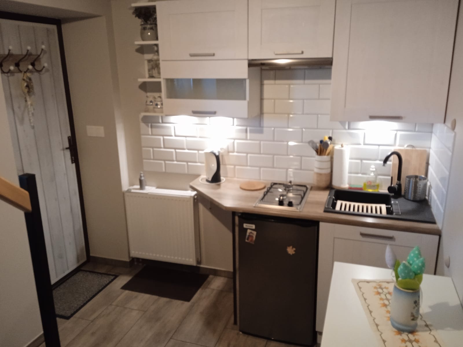 Niewielka kuchnia w apartamencie wakacyjnym z białymi szafkami, ceglanymi płytkami na ścianie i małą lodówką pod blatem, widok na wejście z wieszakami.