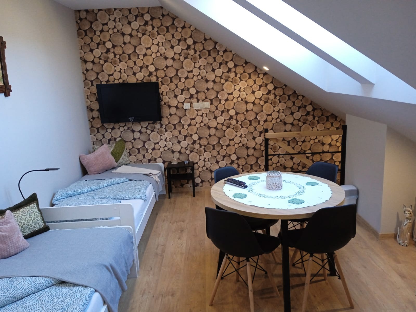 Pokój w pensjonacie z dwoma łóżkami, okrągłym stołem z czarnymi krzesłami i ścianą wyłożoną plastrami drewna, pod skosami dachu z oknami.