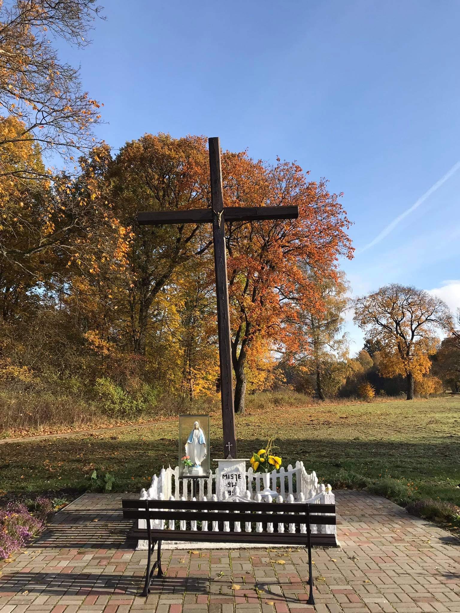 Drewniany krzyż na tle jesiennego krajobrazu, z figurką Matki Boskiej i kwiatami, otoczony niskim białym płotkiem.