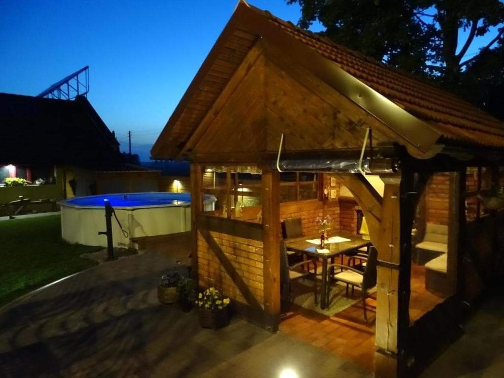 Wieczorny widok na oświetlony basen ogrodowy i drewnianą altanę z ceglanymi ścianami, nakryty stół wewnątrz.