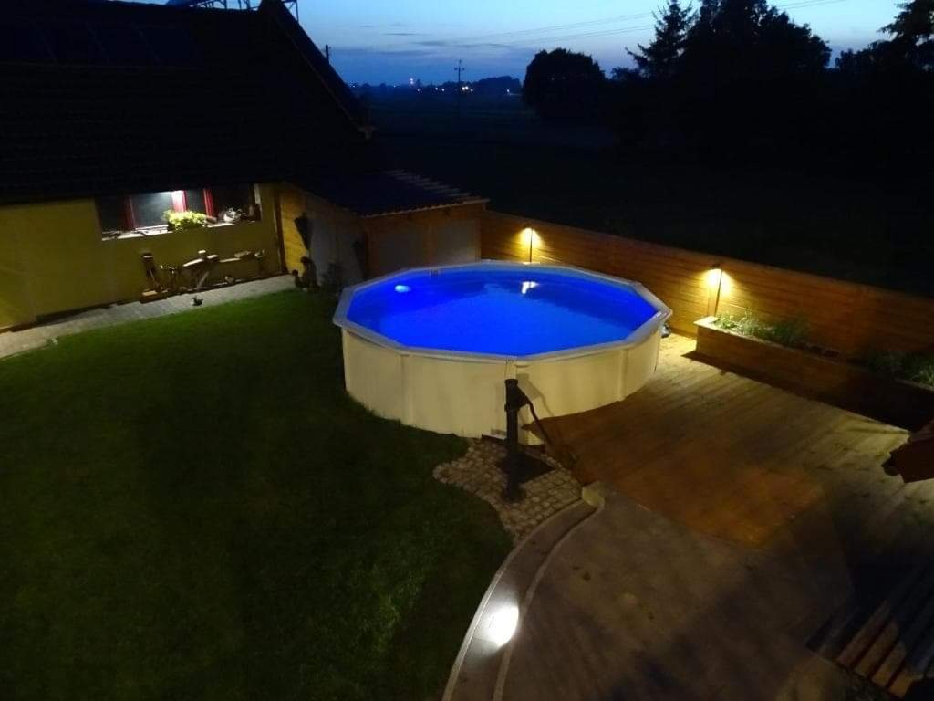 Podświetlony, okrągły basen ogrodowy z białym obramowaniem i niebieską wodą, otoczony drewnianym tarasem i trawnikiem, oświetlony lampami wzdłuż ogrodzenia, widok wieczorny.