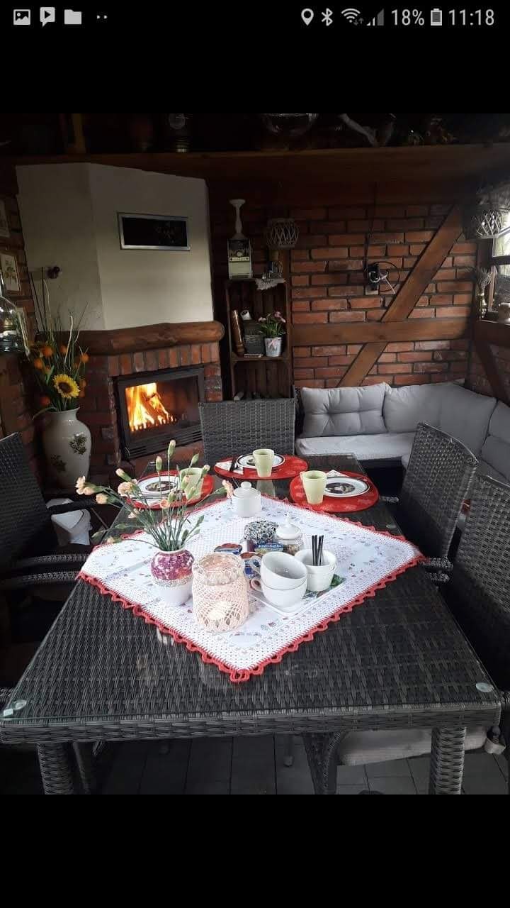 Zadaszony taras z ceglanym kominkiem, nakrytym stołem z wikliny, kwiatami w wazonie i filiżankami.