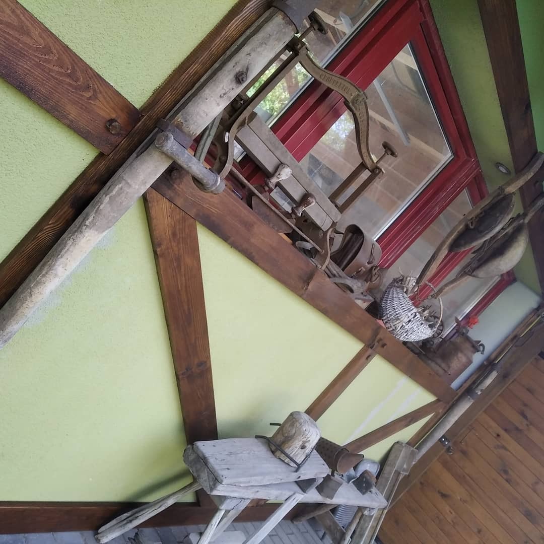 Dekoracyjna ekspozycja starych narzędzi rolniczych i przedmiotów gospodarstwa domowego pod dachem z drewnianą konstrukcją i zieloną ścianą, widoczne okno z czerwoną ramą.