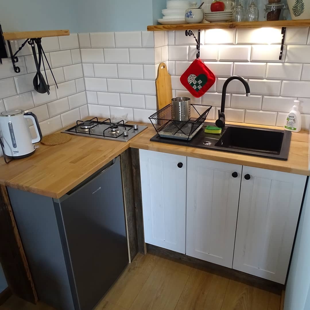 Mała, funkcjonalna kuchnia z białymi płytkami, drewnianym blatem i czarnym zlewem, wyposażona w kuchenkę gazową, małą lodówkę i białe szafki z czarnymi gałkami.