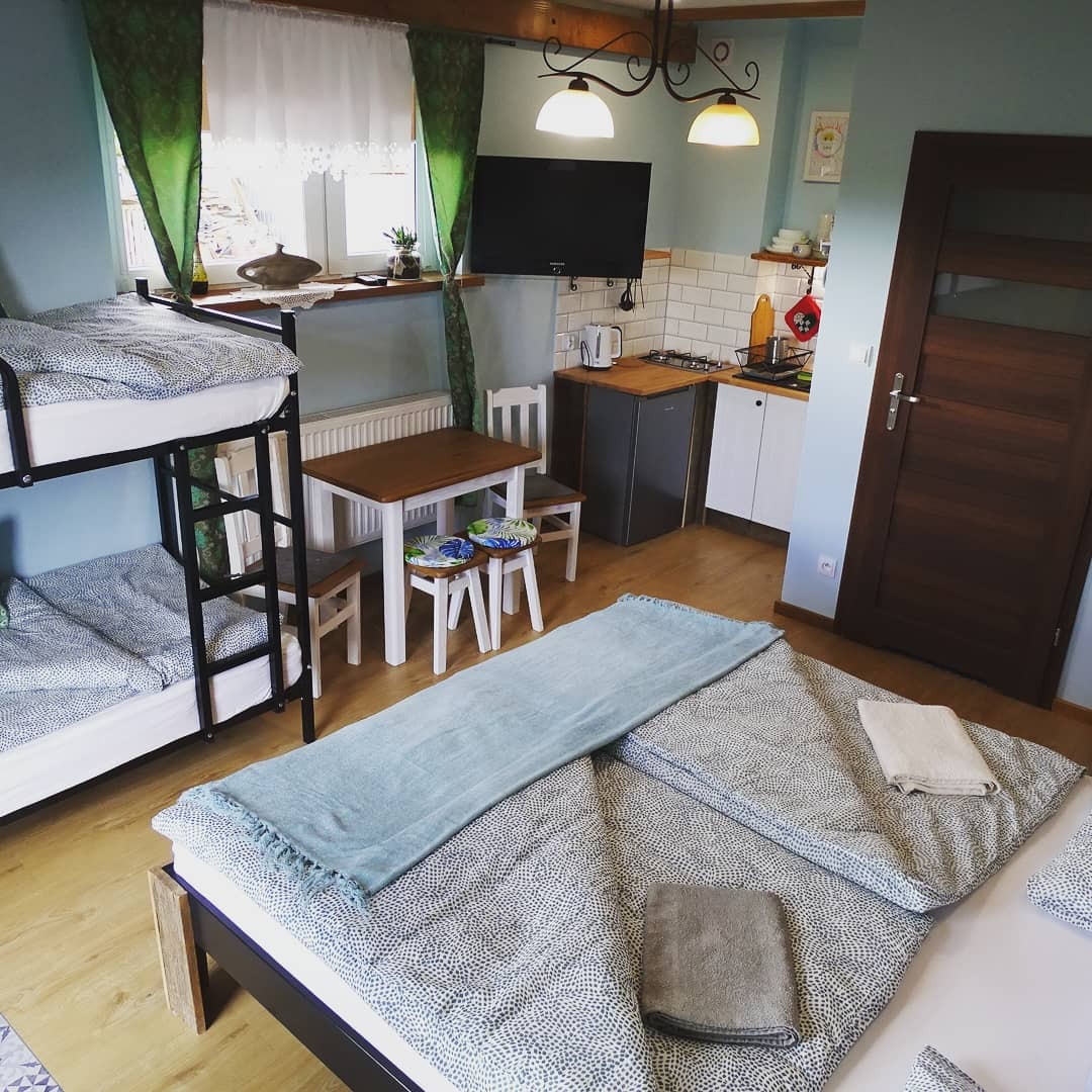 Pokój wieloosobowy z łóżkiem piętrowym, podwójnym łóżkiem i aneksem kuchennym, oświetlony wiszącymi lampami i naturalnym światłem z okna z zielonymi zasłonami.