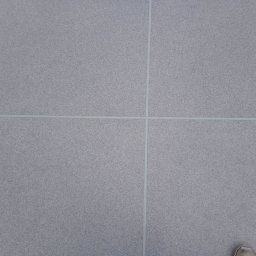 MIR FLOR Mirosław Zarzecki - Szara, drobnoziarnista powierzchnia podłogi podzielona jasnymi fugami na cztery równe kwadraty, fragment buta w prawym dolnym rogu.