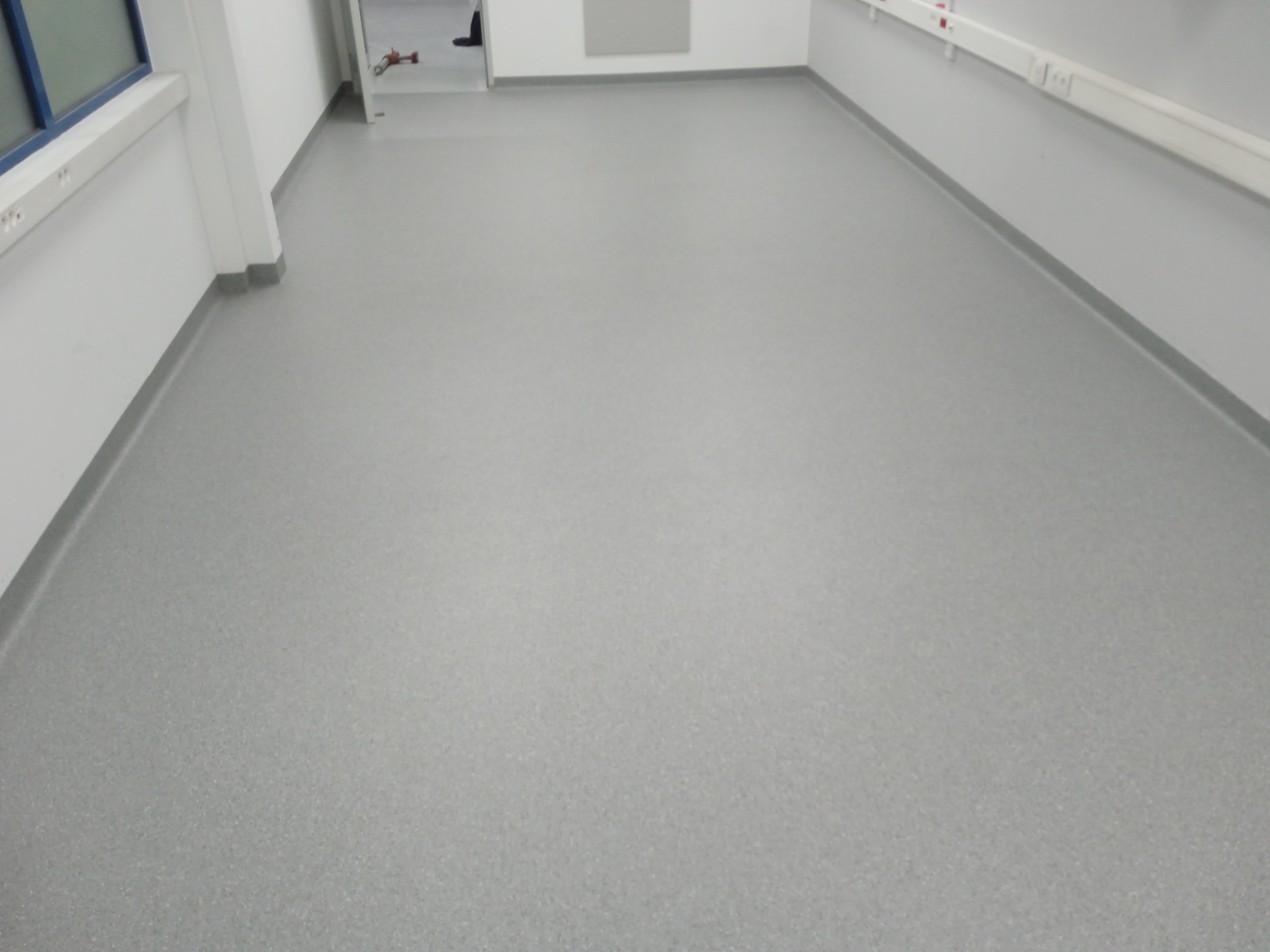 Szare, jednolite wykończenie podłogi w jasnym pomieszczeniu z białymi ścianami i listwami przypodłogowymi, widoczne okno i fragment drzwi w tle.