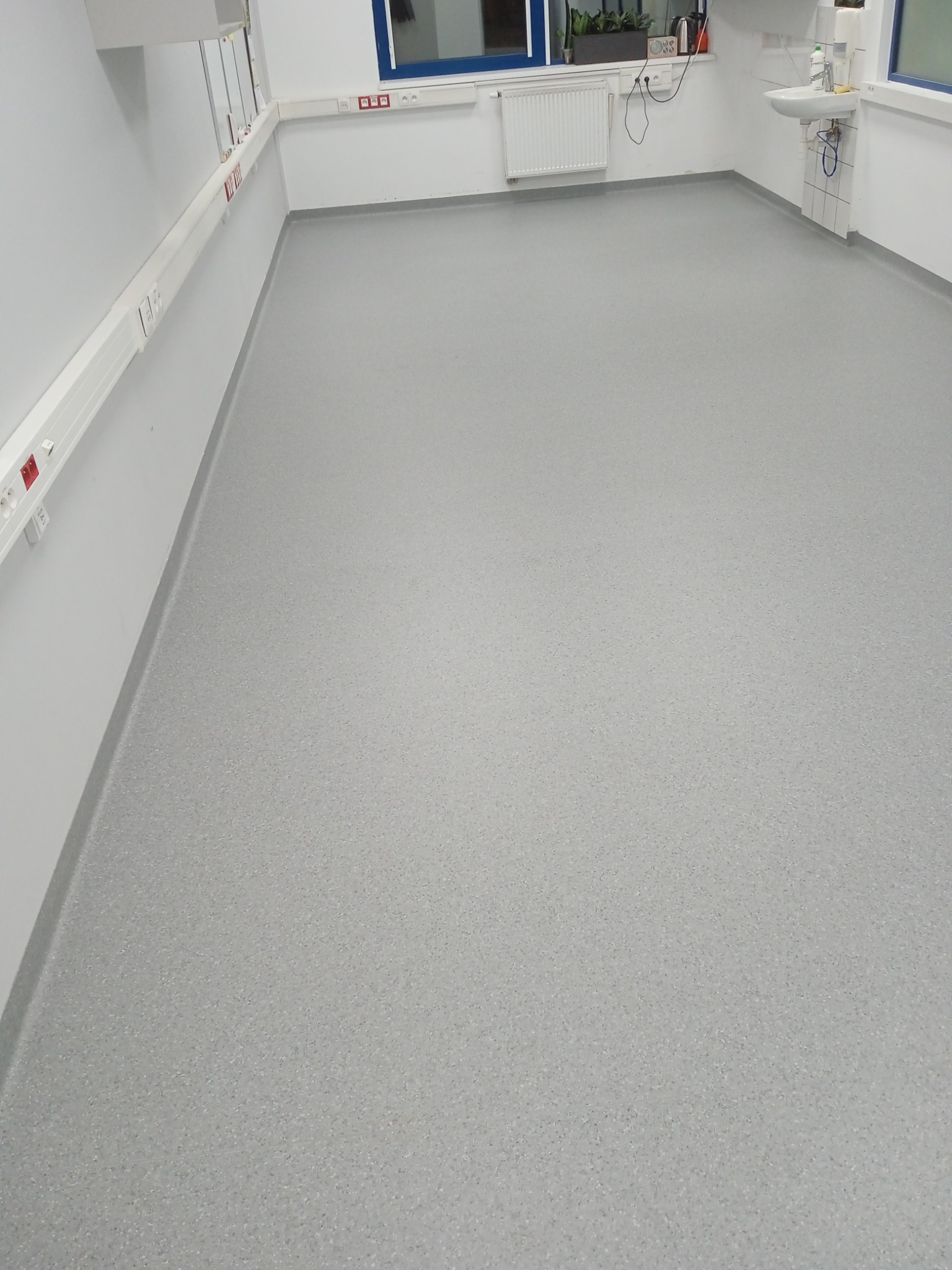 Szara, gładka podłoga w jasnym pomieszczeniu z białymi ścianami, grzejnikiem i umywalką przy oknie.