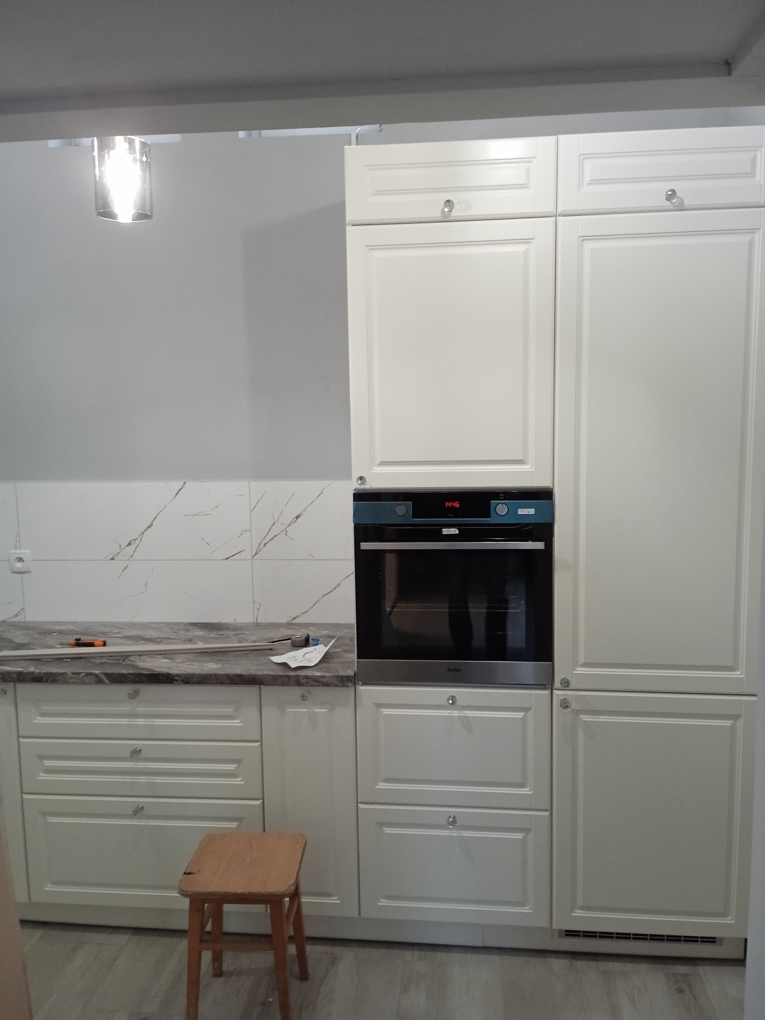 Jasna zabudowa kuchenna z piekarnikiem i marmurkowym blatem, na którym leżą narzędzia. W tle płytki imitujące marmur. Mały drewniany stołek stoi przed szafkami.