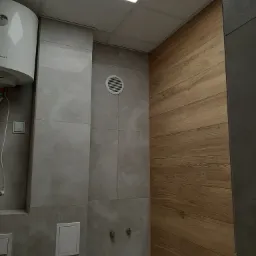 Wykończona łazienka z szarymi płytkami imitującymi kamień, drewnopodobną okładziną ścienną, białym bojlerem i wentylacją.