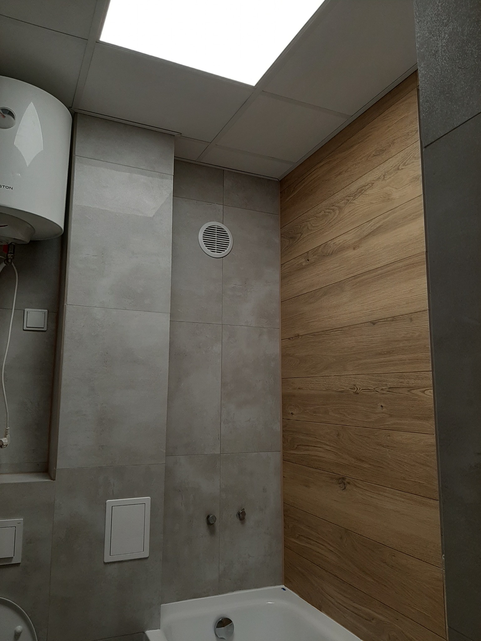 Wykończona łazienka z szarymi płytkami imitującymi kamień, drewnopodobną okładziną ścienną, białym bojlerem i wentylacją.