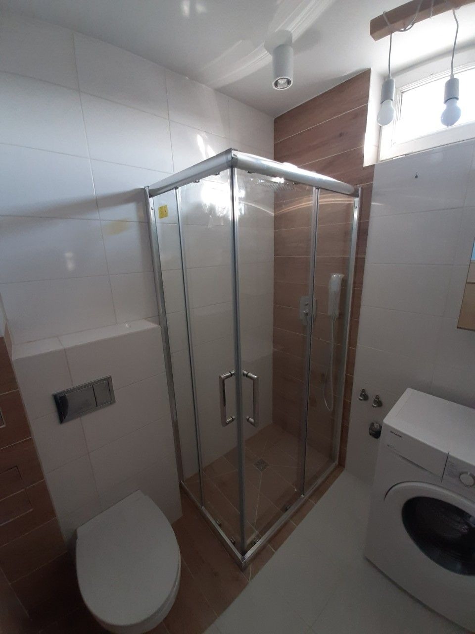 Wyremontowana łazienka z kabiną prysznicową z przezroczystego szkła, białą toaletą wiszącą i pralką. Ściany wyłożone białymi płytkami z brązowymi akcentami.