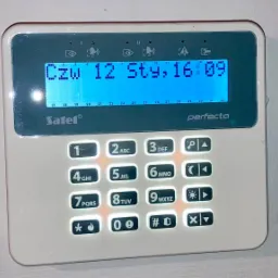 Biały panel sterowania alarmem Satel Perfecta z podświetlanym ekranem LCD wskazującym dzień tygodnia, datę i godzinę, przyciski numeryczne podświetlone pomarańczowym światłem, zamontowany na białej...
