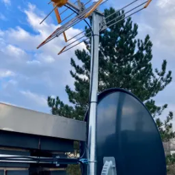 Antena telewizyjna Yagi-Uda z pomarańczowymi elementami i czarna antena satelitarna zamontowane na kominie z ciemnej cegły, widok z dołu na tle błękitnego nieba z chmurami i zielonego drzewa.