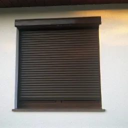 Ciemnobrązowa osłona okienna zewnętrzna, całkowicie zasłonięta, na białej fasadzie budynku z widocznym fragmentem dachu.