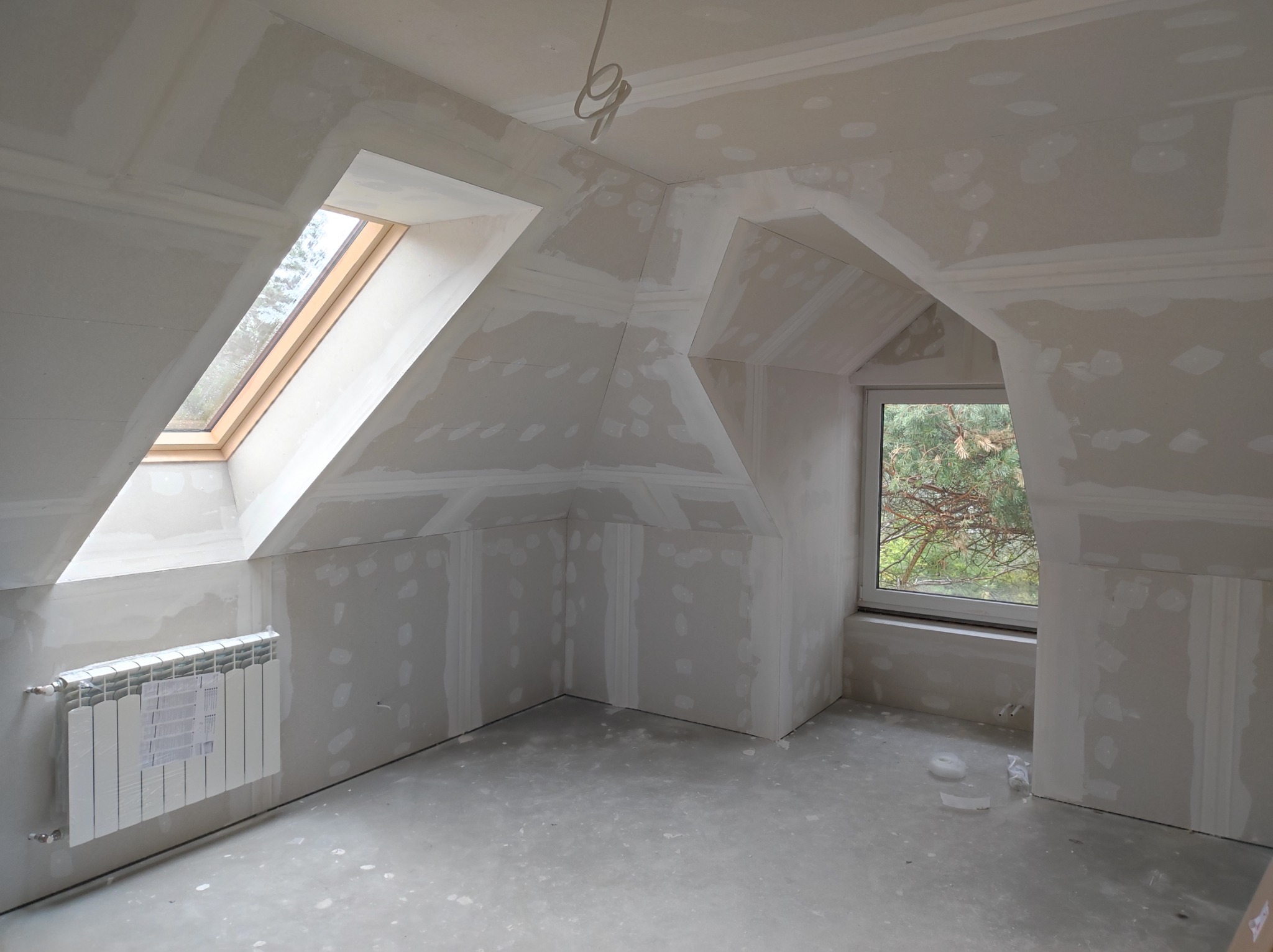 Poddasze w trakcie wykańczania: ściany i sufit z płyt kartonowo-gipsowych po szpachlowaniu, widoczne okno dachowe i grzejnik z folią ochronną.