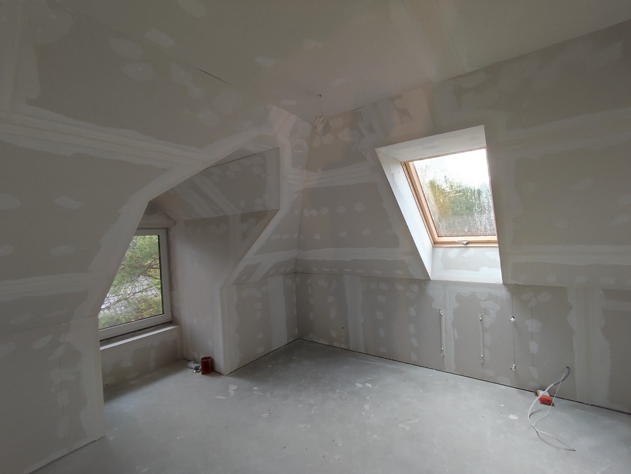 Poddasze po szpachlowaniu, widoczne okna dachowe i okno w ścianie szczytowej, ściany przygotowane do malowania, podłoga zabezpieczona folią.