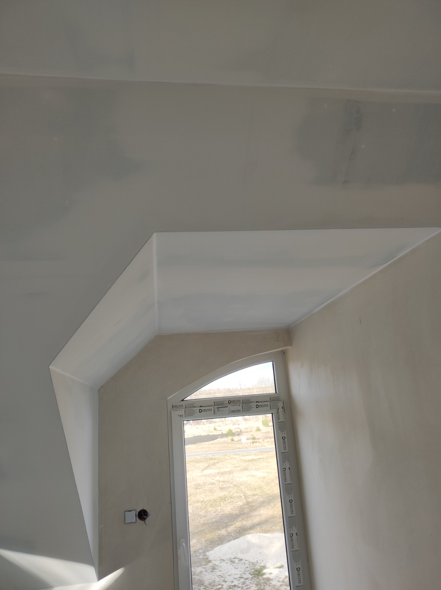 Poddasze ze ścianą i sufitem po szpachlowaniu, widoczne ślady po łatach, okno dachowe z widokiem na zewnątrz, przygotowane do malowania.