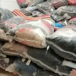 Stos zapakowanych w folię worków z odzieżą, widoczne etykiety na niektórych workach, ułożone w rogu pomieszczenia.