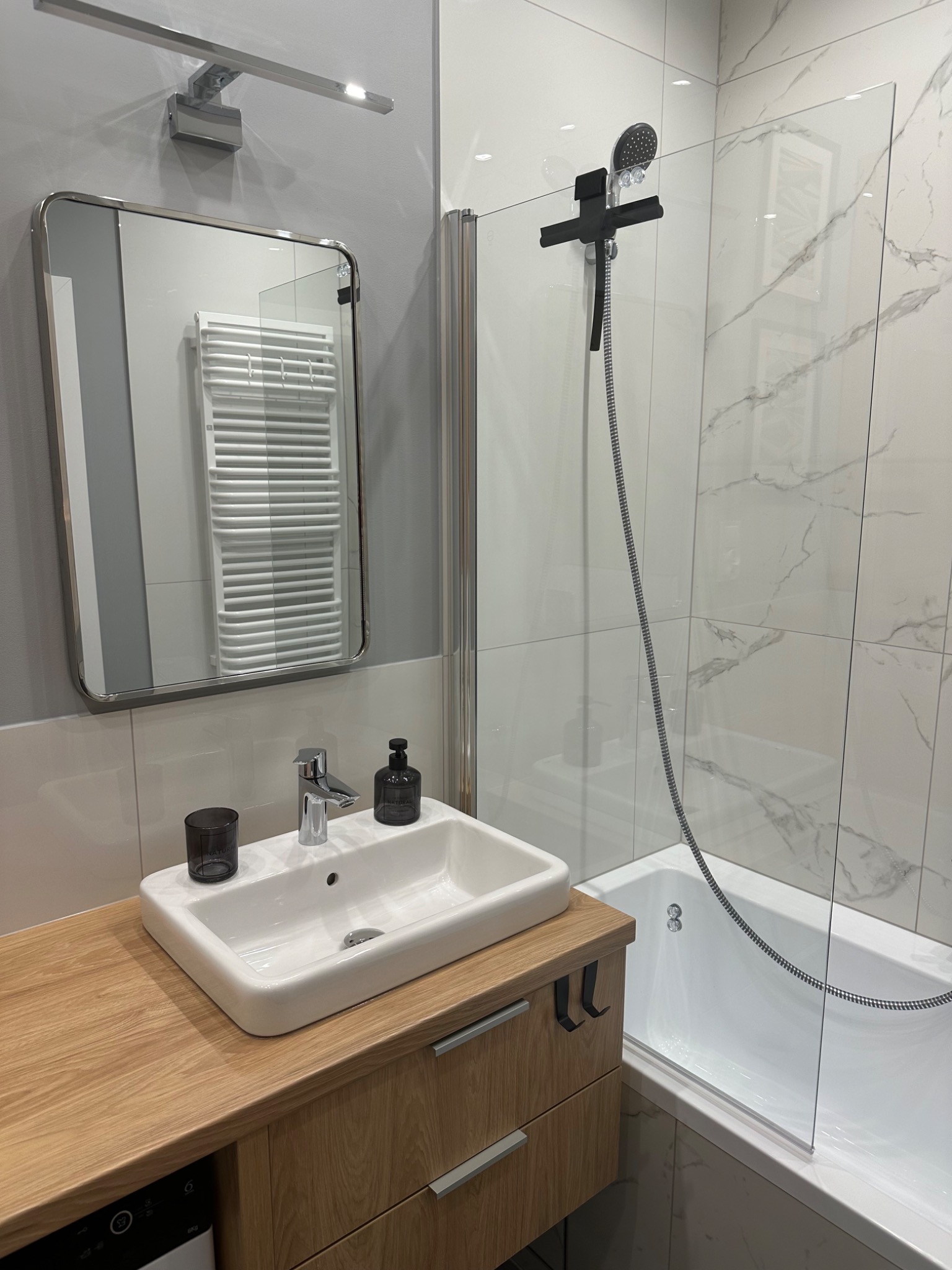 Nowoczesna łazienka z umywalką nablatową na drewnianej szafce, kabiną prysznicową ze szklanym panelem i baterią w kolorze czarnym oraz lustrem z podświetleniem LED odbijającym grzejnik drabinkowy.