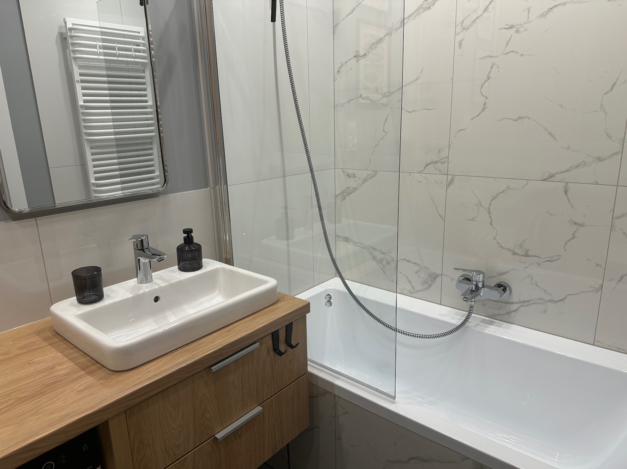 Nowoczesna łazienka z białą armaturą, drewnianą szafką pod umywalką i marmurowymi płytkami ściennymi.