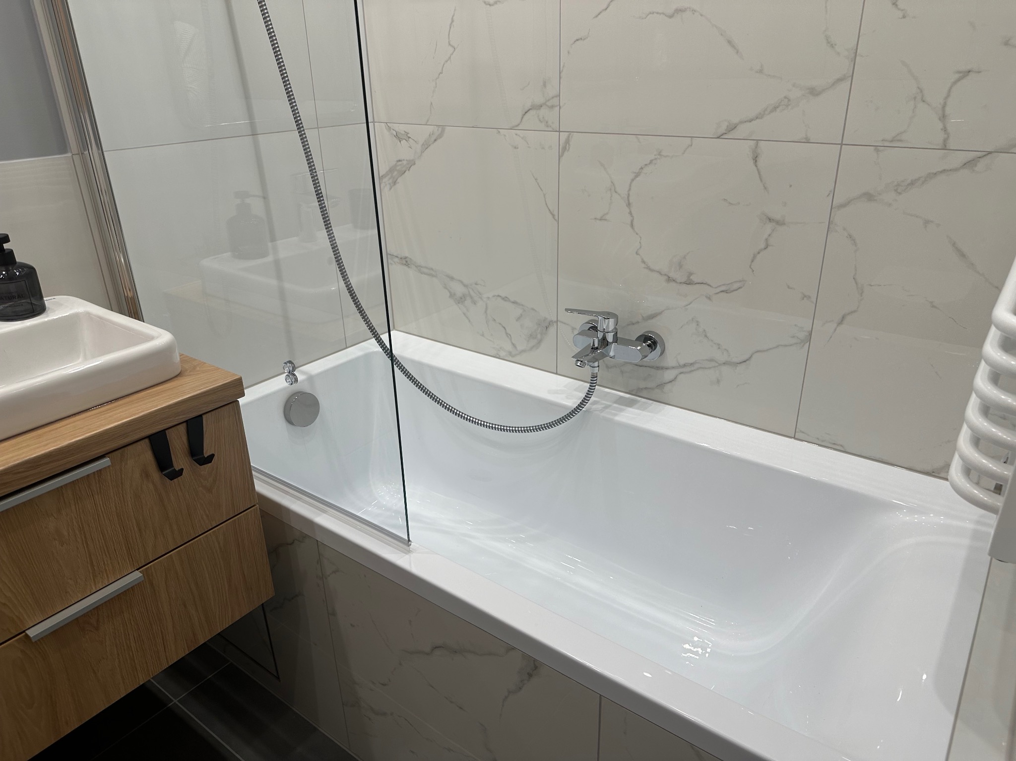 Biała wanna z chromowaną baterią i szklanym parawanem, umywalka z szafką z drewnopodobnymi frontami, marmurowe płytki na ścianach, nowoczesna łazienka.