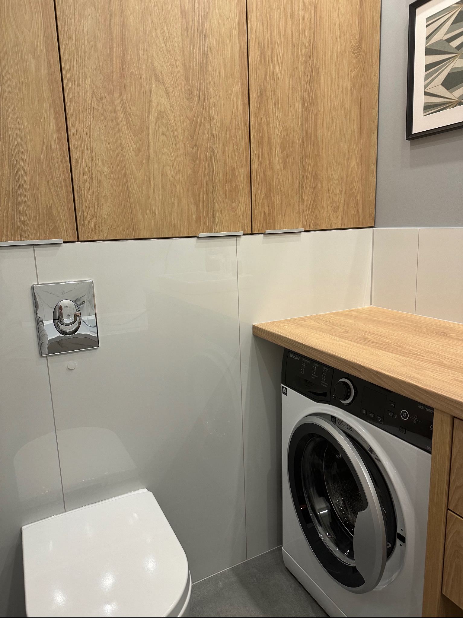 Nowoczesna łazienka z drewnianymi szafkami nad toaletą i pralką Whirlpool pod drewnianym blatem, chromowany przycisk spłuczki na białej ścianie, minimalistyczny obrazek na szarej ścianie w tle.